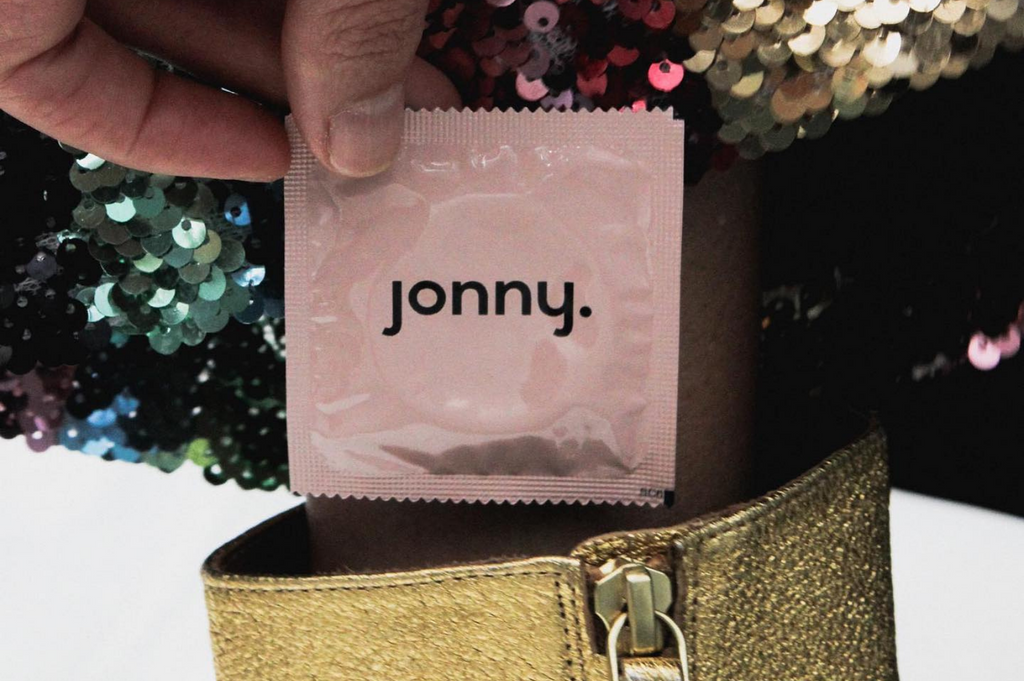 "What is safe sex?" In this image, somebody wearing a sequence pant in putting a pink Jonny condom into a gold boot.