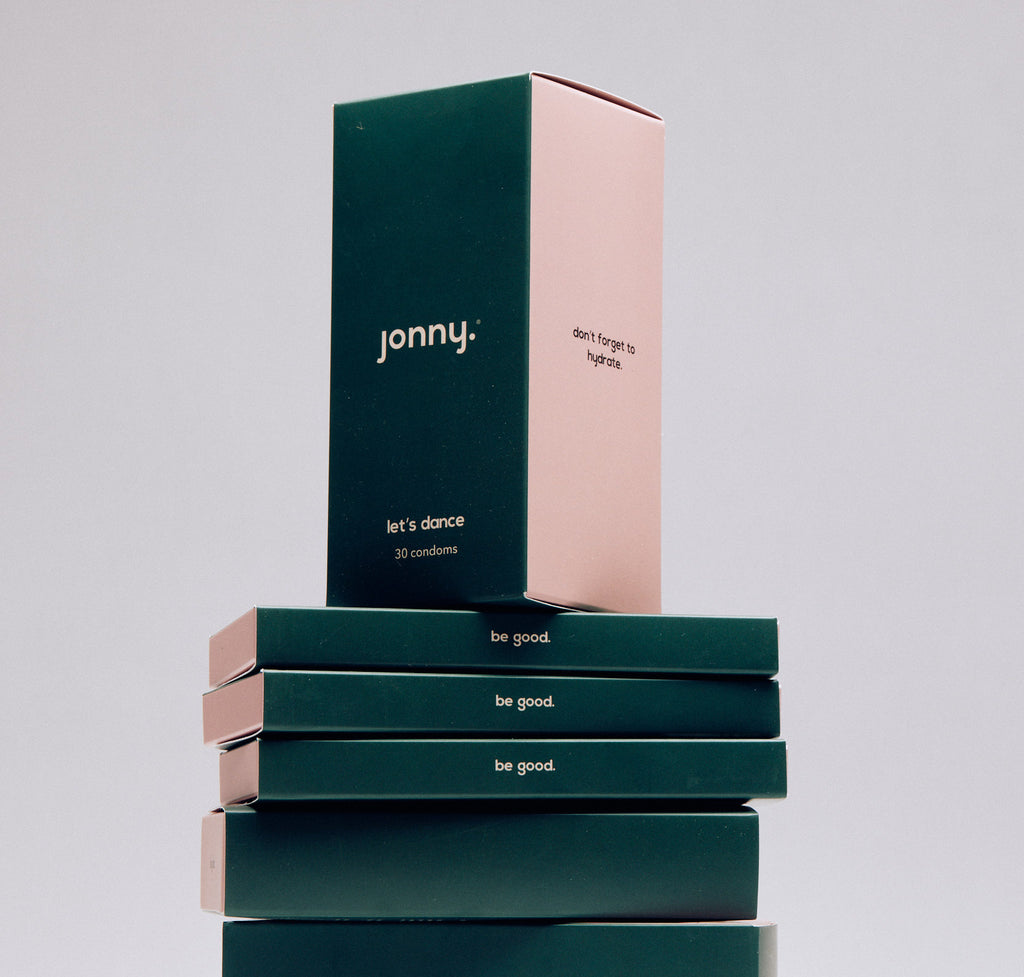 Stacked Jonny condom boxes with eco-friendly packaging, the top box in pink with hydration reminder and the rest in brand's signature dark green with 'be good' slogan, promoting sexual health and sustainability.