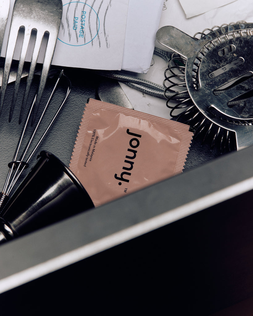 A Jonny brand condom packet in a soft pink hue, casually placed among everyday items such as cutlery and hair styling tools, suggesting a natural integration of safe sex practices into daily life.
